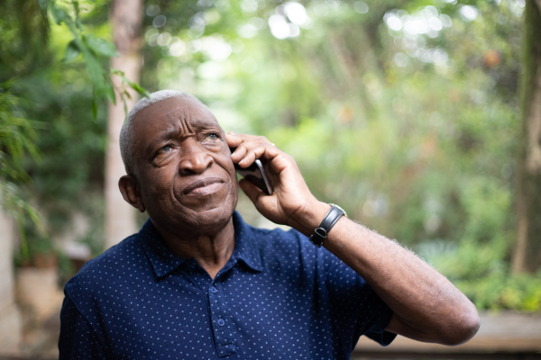 An older man with short grey hair, wearing a navy blue, short-sleeved, button-up shirt with small white dots, stands outdoors among greenery. He holds a mobile phone to his ear and looks up thoughtfully with a slight frown.