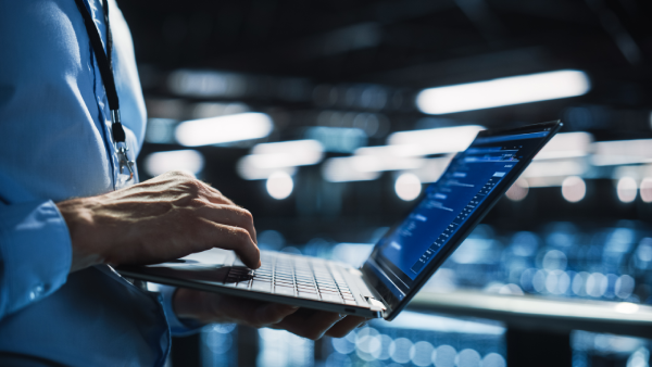 A person in a blue shirt uses a laptop, typing with one hand. The laptop screen displays lines of code. The background is blurred, with bright lights and a modern, industrial setting, suggesting a tech or data center environment.