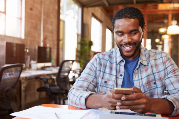 A man with short hair and a beard, wearing a checked shirt over a blue T-shirt, sits at a desk in a modern open-plan office. He smiles whilst looking at his mobile phone. Papers and a tablet are on the desk; empty chairs and computers are behind him.