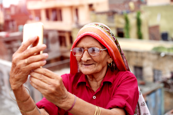 An elderly woman wearing glasses, a red sari blouse, and a colorful dupatta smiles while taking a selfie with a smartphone outdoors, with blurred buildings in the background. She appears happy and engaged with the device.