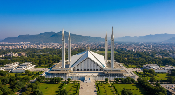 Aerial view of the Faisal Mosque in Islamabad, Pakistan, showing its white triangular roof and four tall minarets, surrounded by greenery, with the cityscape and Margalla Hills in the background under a clear blue sky.