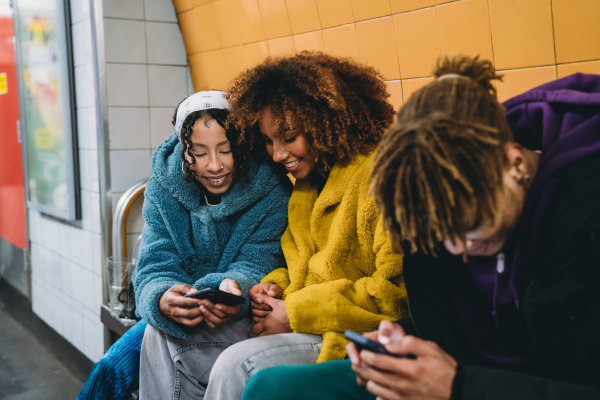 Three young people in colourful jackets sit on a bench in an underground station, looking at their mobiles and smiling. The wall behind them is tiled in yellow and white. The mood is cheerful and relaxed.