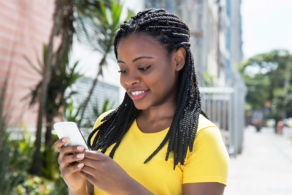 A smiling Black woman with dark braided hair wears a bright yellow top whilst looking down at a mobile phone in her hands. She stands outdoors on a sunny day with blurred trees and a city street in the background.