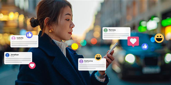 A woman stands on a city street at dusk, looking at her phone. Around her, digital icons of likes, hearts, and smileys, along with comment bubbles labeled Isabella, Jonathan, Katherine, and Ramsey, represent social media interactions. Cars and lights blur in the background.