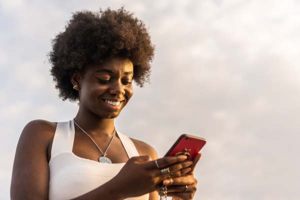 A young woman with a natural afro hairstyle smiles whilst looking at her red mobile phone. She wears a white sleeveless top, a pendant necklace, and rings. The sky is partly cloudy in the background. The lighting is warm and natural.