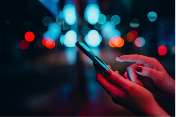 A close-up of hands with red-painted nails using a mobile phone at night. The background is blurred, featuring colourful blue and red lights, creating a bokeh effect that suggests an urban, city environment.