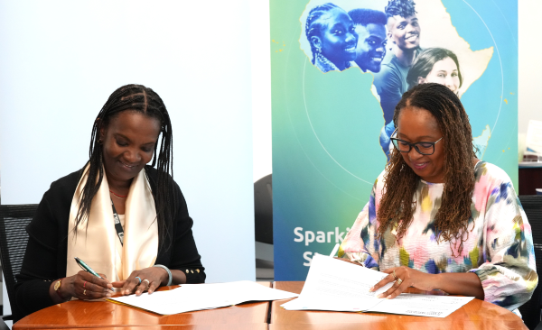 Two women sit at a wooden table signing documents. Both are smiling and focused on their papers. Behind them is a banner featuring outlined faces, an African map, and the words “Sparkle SA”. The setting appears bright and formal.