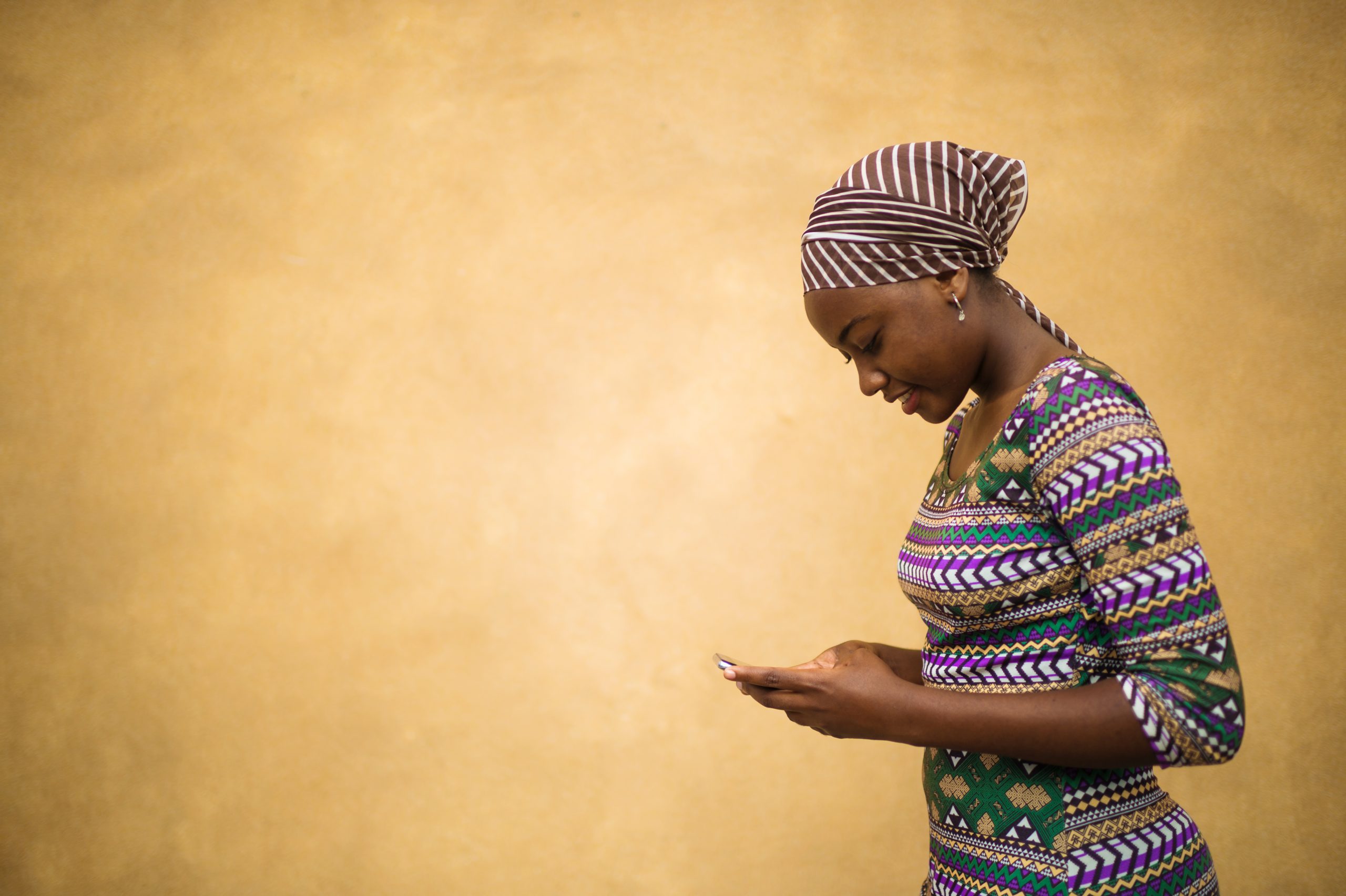 Side view of a young African girl dressed in traditional clothing and a headscarf standing against a plain brown yellow warm background communicating on her mobile phone in Dar es Salaam East Africa