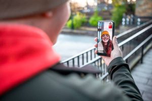 Person video calling someone outdoors, showing a smiling woman on the phone screen wearing a yellow hat. using phone for video call and connectivity
