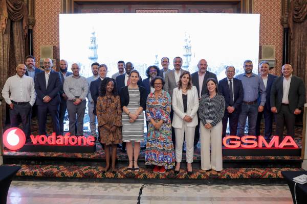 A group of 22 people, from GSMA and Vodaphone stand on a stage in two rows. Behind them is a large, bright screen showing minarets. The Vodafone and GSMA logos are displayed in red on the front of the stage.