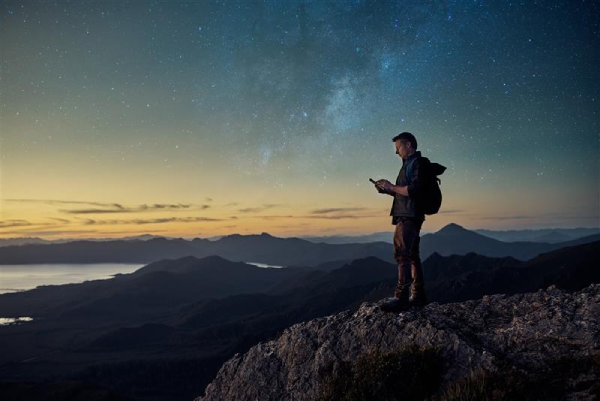 A person with a backpack stands on a rocky cliff at dusk, looking at a smartphone. Mountains and a large lake stretch out below, while a starry sky with the Milky Way glows above the horizon. The scene captures a sense of adventure and solitude.