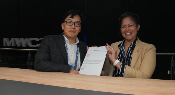 Two people sit at a wooden table, smiling and holding up a document together. The man on the left wears glasses and a dark checkered jacket; the woman on the right wears a tan blazer and dark scarf. The background features the letters "MWC" in a dimly lit setting.