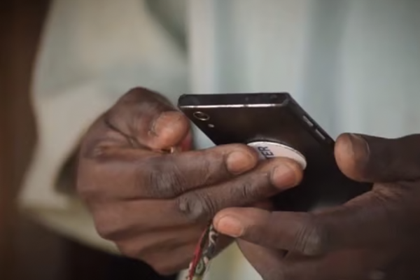 Close-up of a person’s hands holding and using a black mobile phone with a pop socket. The phone’s screen is not visible. The person wears a light-coloured long-sleeve garment, and the background is blurred.