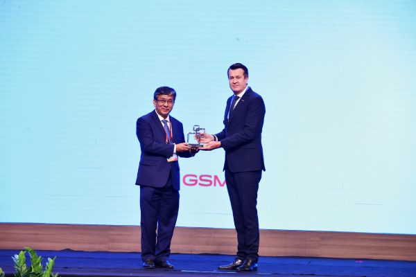 Two men in dark suits stand on a stage as one hands a small, geometric silver trophy to the other. They are positioned in front of a large, pale blue screen featuring the red letters “GSM”.
