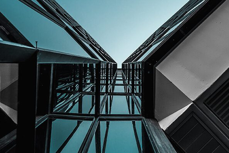 Upward view of a modern glass building with reflective windows. Two sections of the structure converge toward the sky, forming a symmetrical V-shape. The sky is a clear, light blue. The image highlights the sleek, geometric lines and the contrast between glass and matte surfaces.