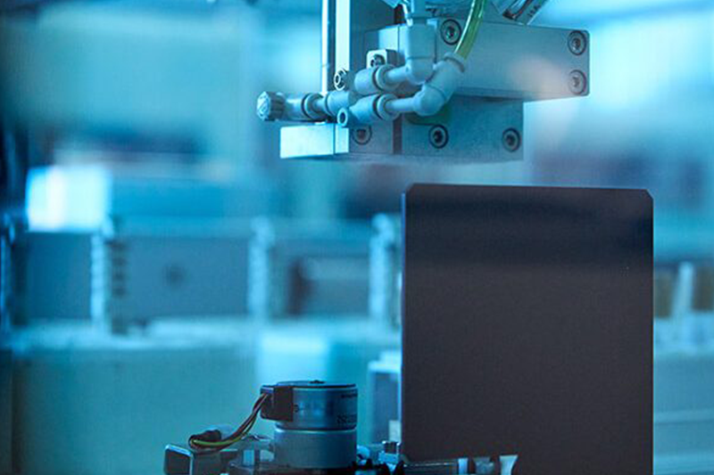 A close-up of a robotic assembly line in a factory, with a mechanical arm positioned above a flat, rectangular object, possibly a smartphone or electronic device. The scene is bathed in blue light, emphasizing the technological and industrial atmosphere.