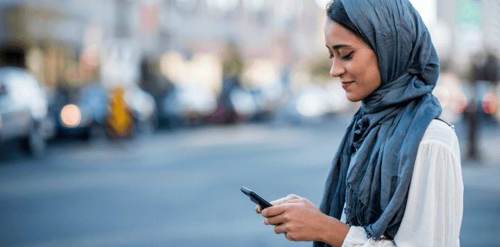 A woman wearing a hijab looks at her smartphone on a busy street, embodying sustainable and responsible business leadership.