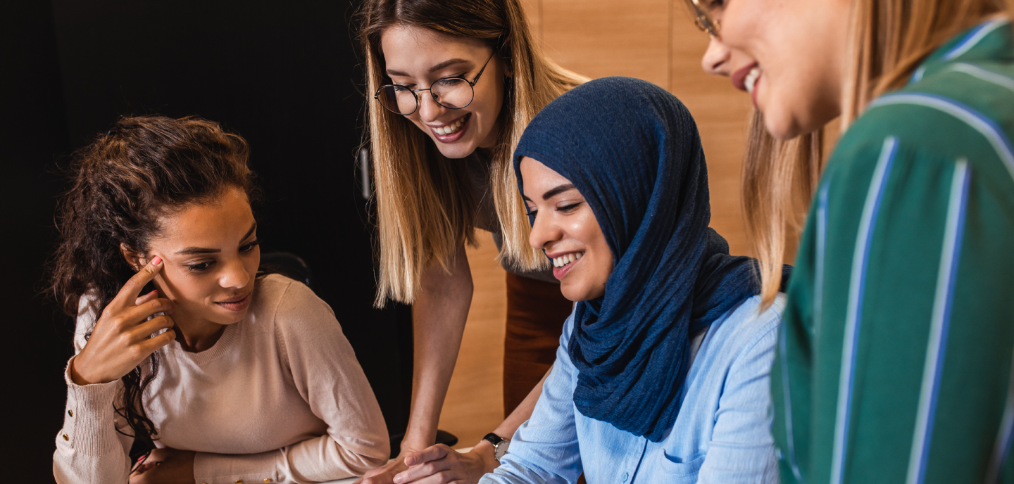 Four women, one wearing a hijab, smiling and engaging in discussion around a table in a brightly-lit room.