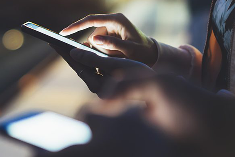 Close-up of two hands using smartphones in dim lighting. A person is tapping on one phone screen, while the other hand holds another phone in the foreground. The screens emit a soft glow, creating a contrast against the dark background.