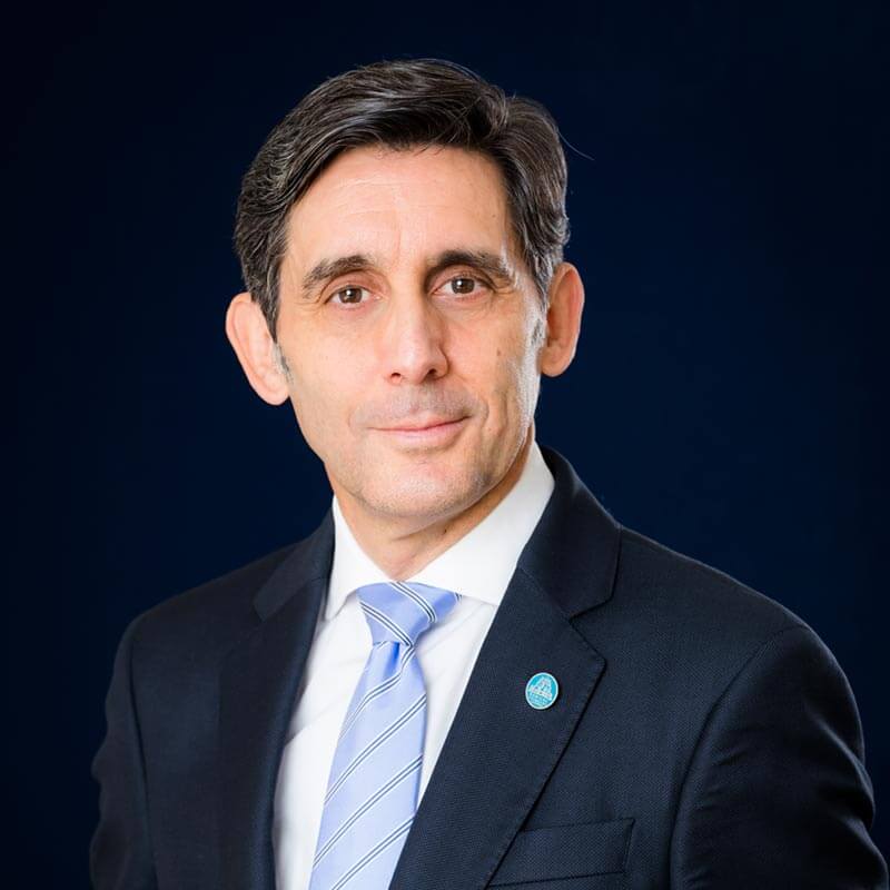 A man with short dark hair is wearing a dark suit, light blue tie, and a small blue lapel pin. He is posing against a dark blue background, looking directly at the camera with a neutral expression.