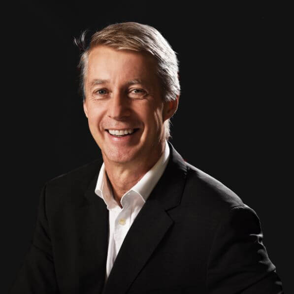 A man with gray hair smiles at the camera. He is wearing a black suit jacket, a white dress shirt with the collar slightly open, and has a dark background behind him.