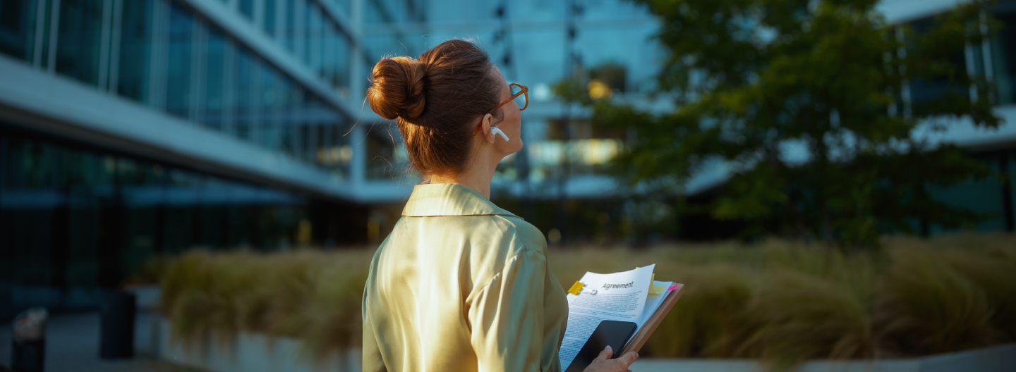 A woman with brown hair in a bun, wearing glasses and a light coat, stands outdoors holding documents and a smartphone. She faces a modern glass office building, reflecting sustainable and responsible business leadership amid plants and trees.