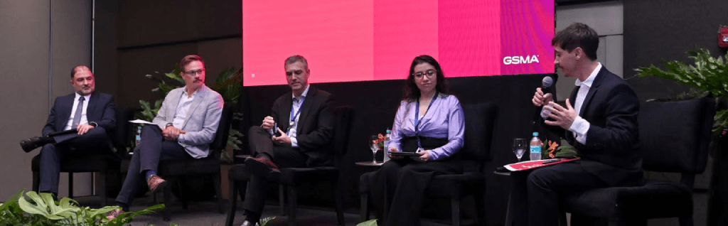 Five panellists in business attire sit on a stage before a GSMA screen to discuss digital inclusion. The group includes four men and one woman, with the person on the far right holding a microphone and gesturing as they speak. Side tables with glasses of water sit between them.