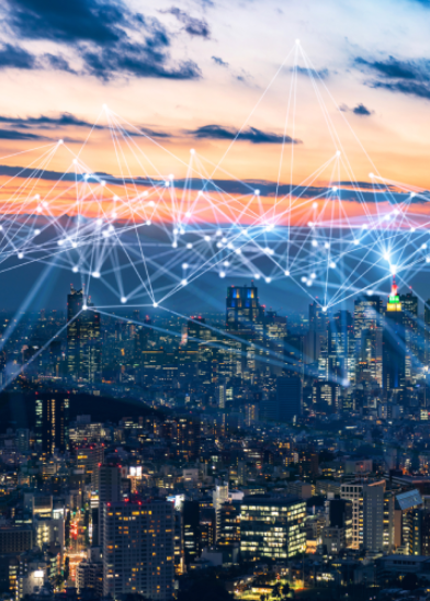 A city skyline at sunset with tall buildings illuminated. Digital white lines and glowing dots are superimposed over the scene, representing a network or smart city connectivity, blending technology with the urban landscape under a colorful sky.