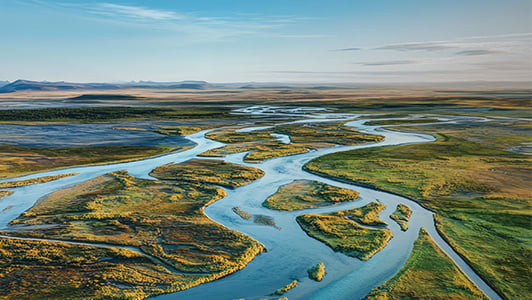 A wide aerial view of a winding river branching into multiple channels across a vast, green landscape. The sky is clear with a few clouds, and distant mountains are visible on the horizon. The scene is bright and serene, showing untouched wilderness.