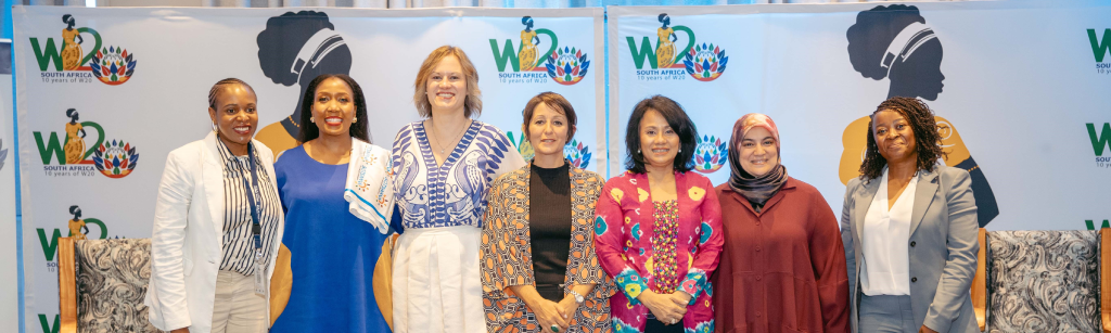 A diverse group of eight women stand side by side, smiling at the camera. They wear a mix of business and traditional attire. Behind them, there are banners featuring the "W20 South Africa" logo and silhouetted profiles of women’s faces.