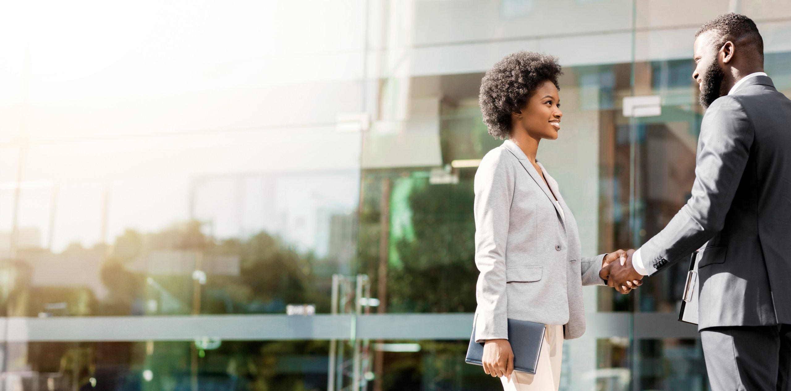 A woman and man in professional business attire stand outside a modern glass building, smiling and shaking hands. The woman holds a tablet, suggesting a discussion about responsible AI policy in today’s bright, positive, and professional atmosphere.