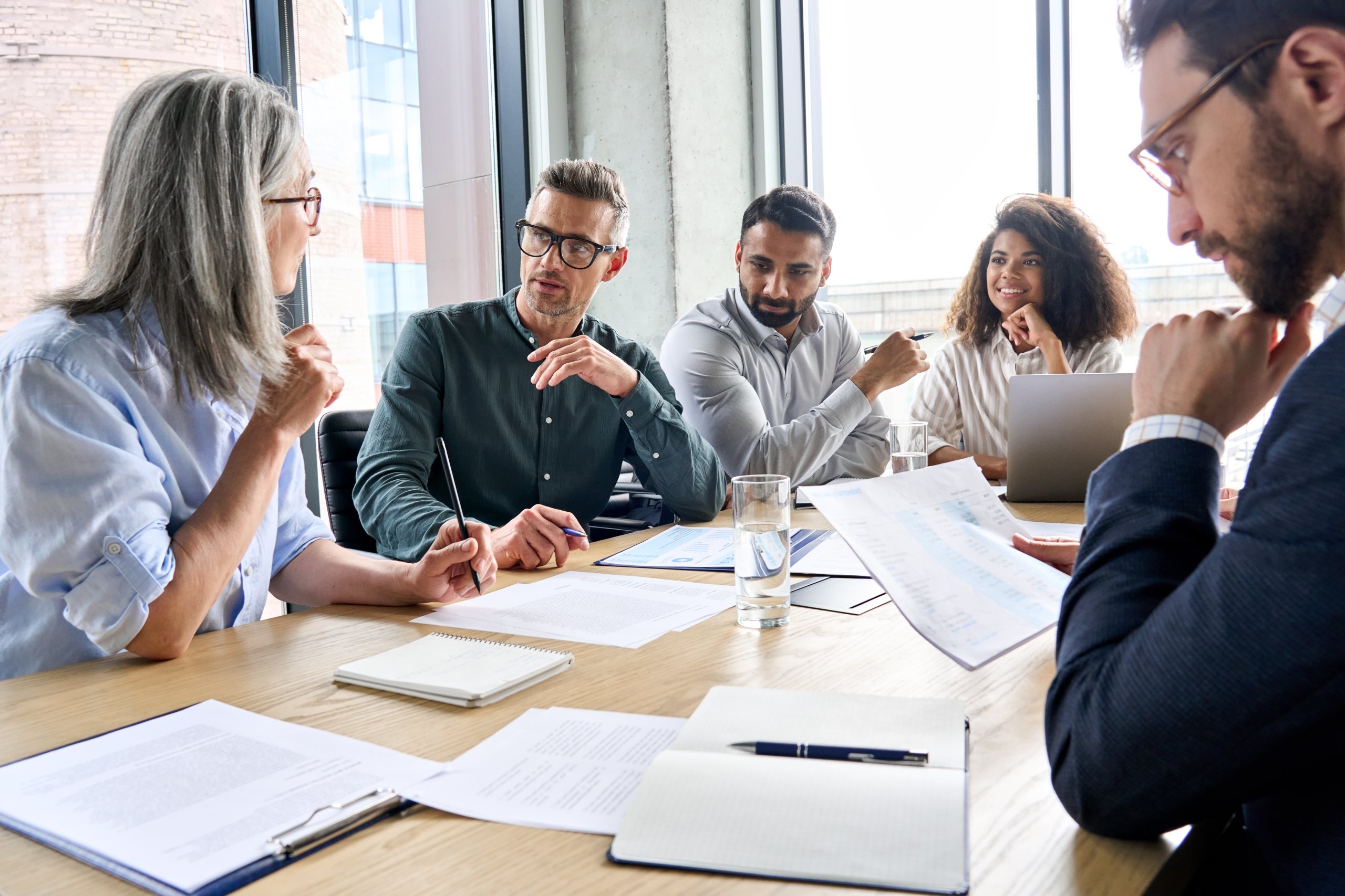 Five people sit around a table in a bright office, engaged in discussion. Papers, notebooks, and a laptop are on the table. One woman gestures while others listen attentively. Large windows let in natural light, creating a collaborative atmosphere.