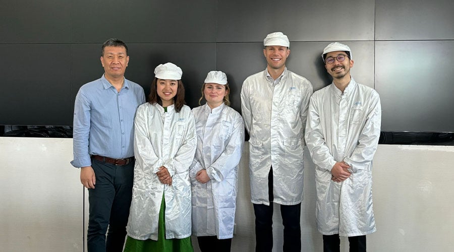 Five people stand indoors in front of a black wall. Four wear white lab coats and caps, while one wears a light blue shirt and dark pants. All are smiling, suggesting a professional or scientific setting focused on Climate Action in China.