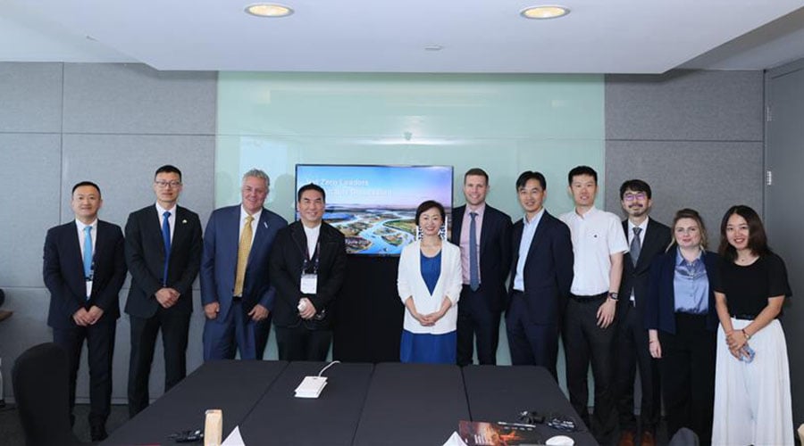 A group of eleven people, diverse in gender and ethnicity, stand indoors in business attire, posing for a photo. Behind them is a wall-mounted screen displaying an aerial image and text related to Climate Action in China.