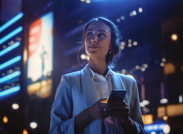A woman in a white blazer stands outside at night, holding a smartphone and looking upward with a slight smile. Neon lights and billboard signs glow in the background, highlighting the modern impact of mobile AI on city life and optimism.