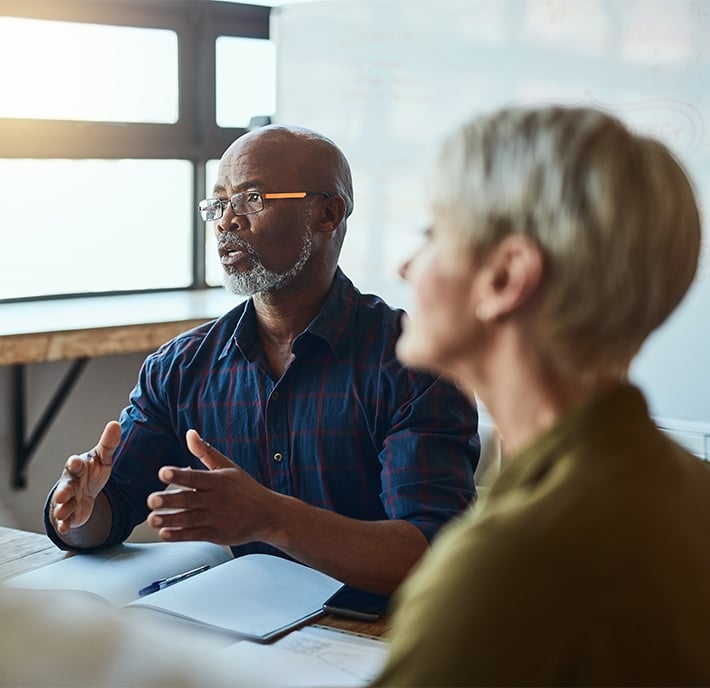 A man with glasses and a grey beard discusses Responsible AI, gesturing with his hands in a meeting room. He sits at a table with an open notebook, facing a woman with short blonde hair. Sunlight streams through large windows in the background.