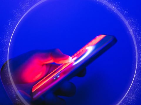A hand illuminated by neon pink and blue light holds and swipes on a smartphone screen, highlighting the digital divide. The deep blue background and glowing circular aura create a futuristic digital effect.