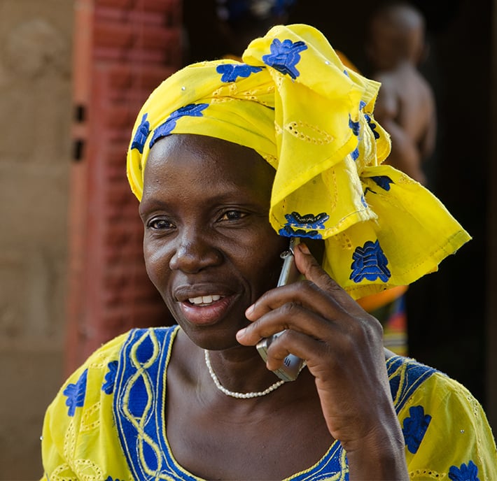 A woman wearing a bright yellow headwrap and dress with blue patterns smiles while talking on a cellphone, highlighting how connectivity helps bridge the digital divide. She wears a pearl necklace and stands outdoors by a reddish wall, another person behind her.
