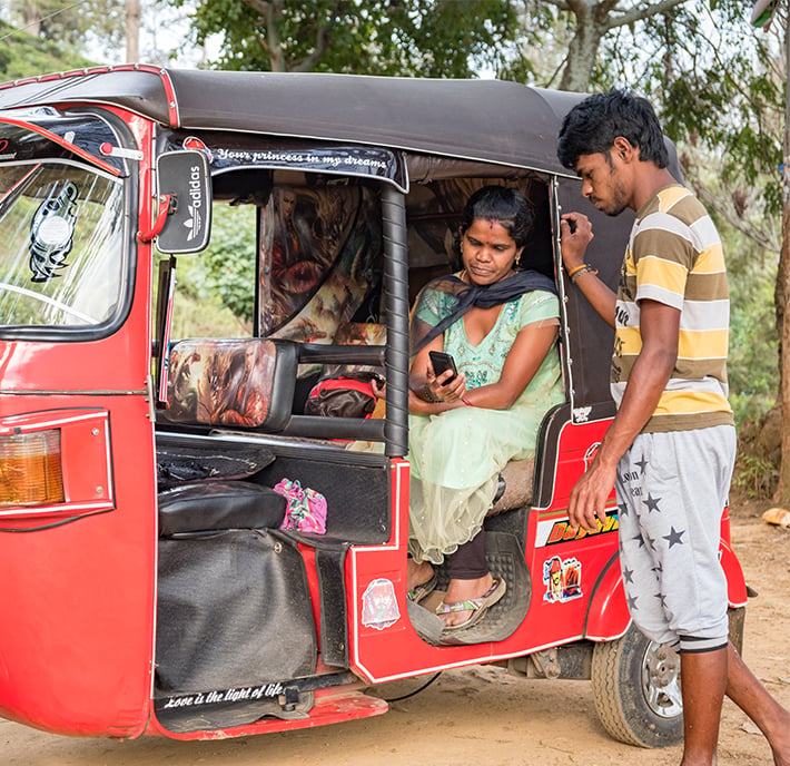 A woman in a green dress sits inside a red auto rickshaw, looking at her smartphone, while a man in a striped shirt stands outside, both focused on the screen—a subtle moment that hints at the digital divide amidst an outdoor setting with trees.