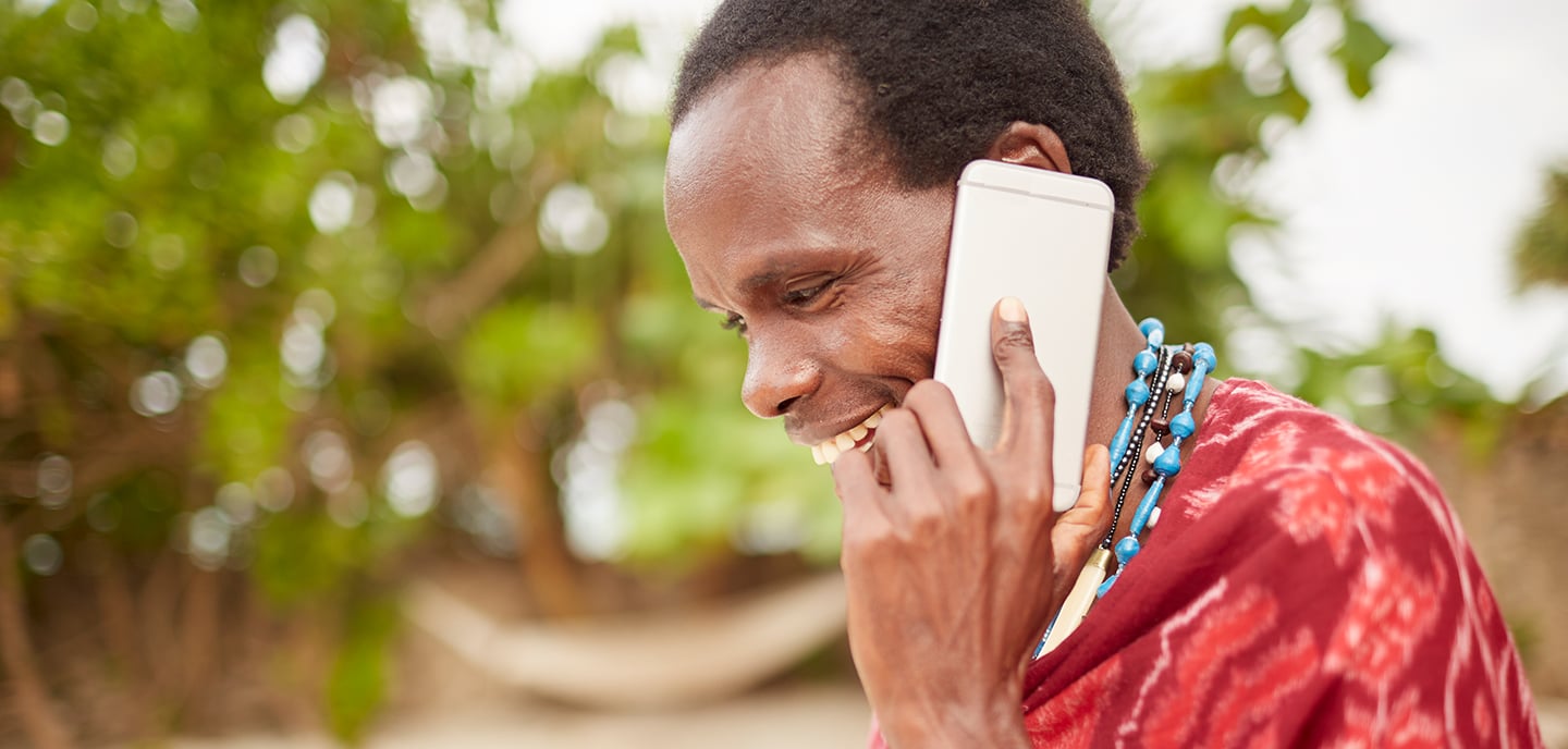 A person wearing a red patterned garment and beaded necklace smiles while holding a smartphone to their ear, bridging the digital divide. Green foliage and a hammock are blurred in the background, suggesting a relaxed, sunny environment.