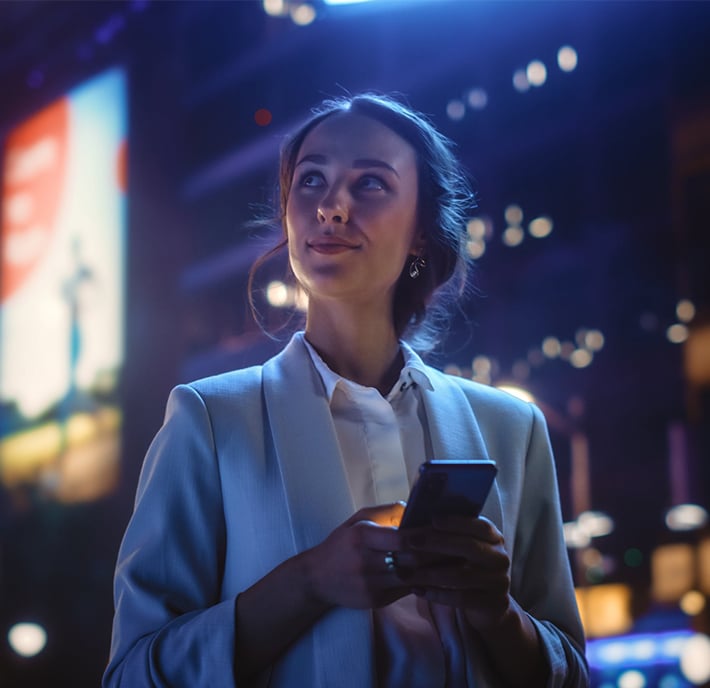 A woman in a light blazer, possibly working in External Affairs, holds a mobile phone and smiles softly whilst looking upward. She stands outdoors at night with blurred city lights and a bright digital hoarding behind her.