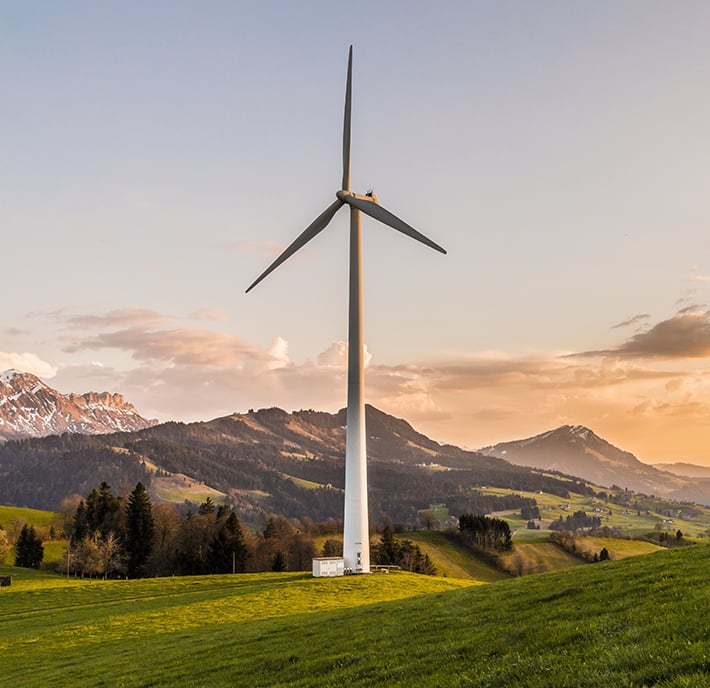 A large wind turbine, part of an External Affairs sustainability initiative, stands on a grassy hill at sunset, surrounded by rolling green fields, scattered trees, and distant snow-capped mountains under a pastel sky with soft clouds.