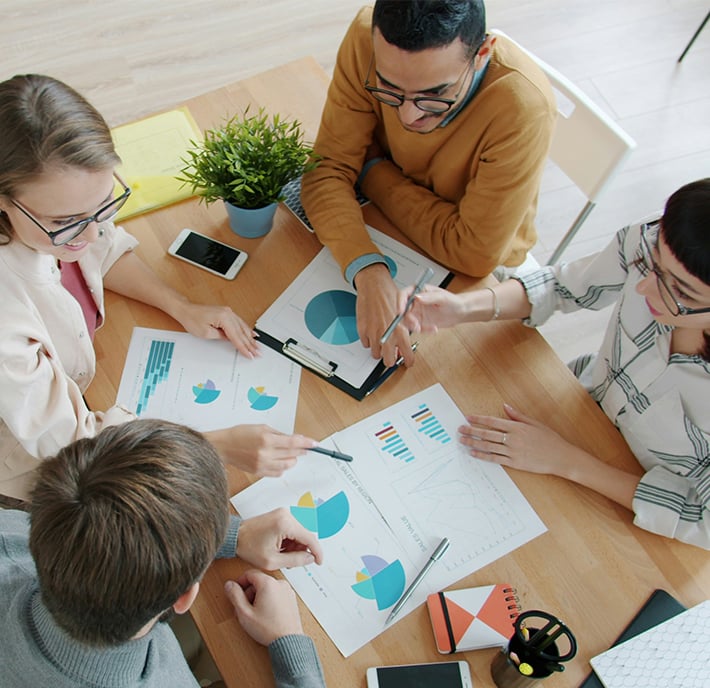 Four people sit around a table reviewing colourful charts and graphs on paper, discussing net zero strategies. Laptops, a mobile phone, a plant, and notebooks are on the wooden table as the group collaborates closely on climate change solutions.