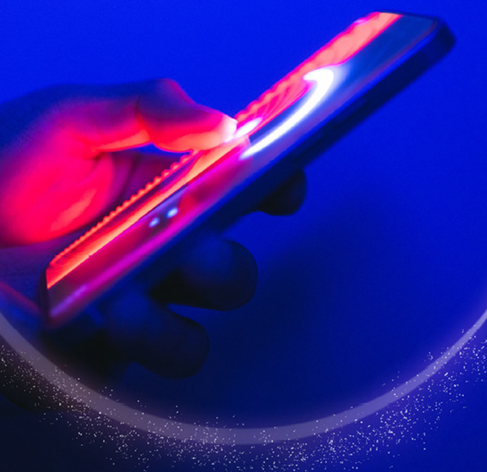 A hand holds a smartphone glowing with vibrant red and blue neon lights. The device’s screen emits a bright white curved line, creating an arc of light and sparkling particles against a deep blue background. The scene feels futuristic and dynamic.