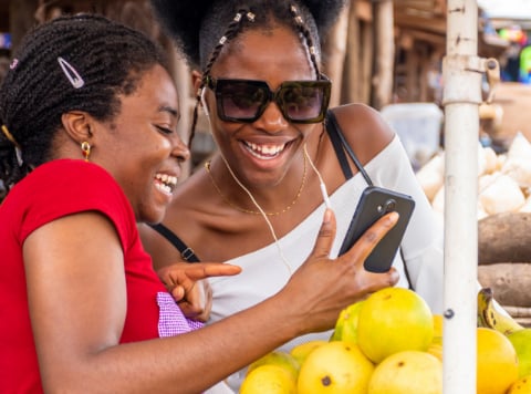 Two women smile and laugh together while looking at a smartphone, bridging the digital divide. One wears a red top, the other a white one with large sunglasses. In the foreground are yellow fruits piled on a market stall, with stalls blurred in the background.