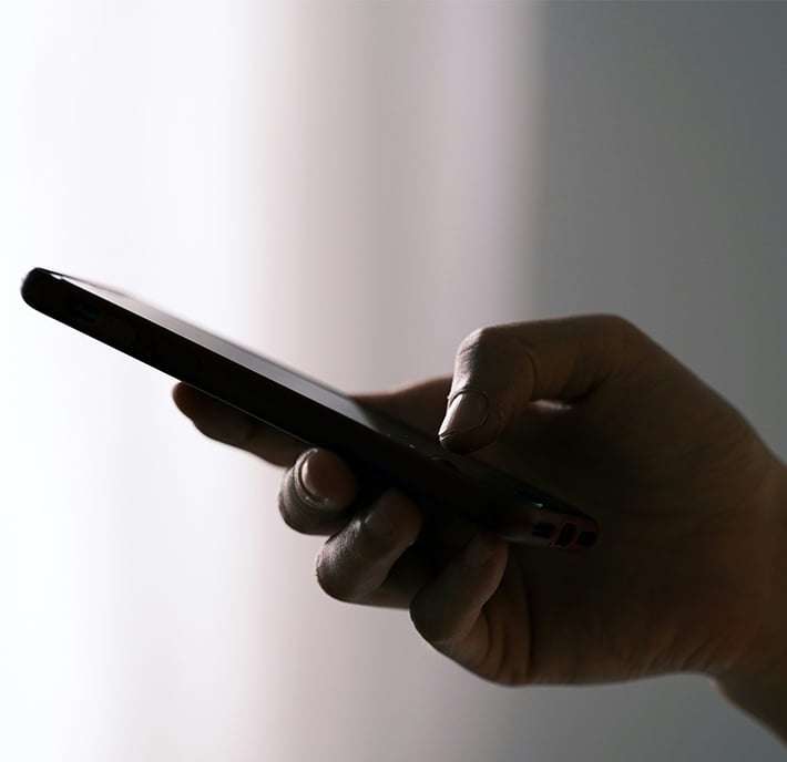 A close-up of a person’s hand holding a smartphone against a softly lit, blurred background. The thumb is touching the screen, suggesting they are interacting with an External Affairs app. The overall lighting is dim and natural.