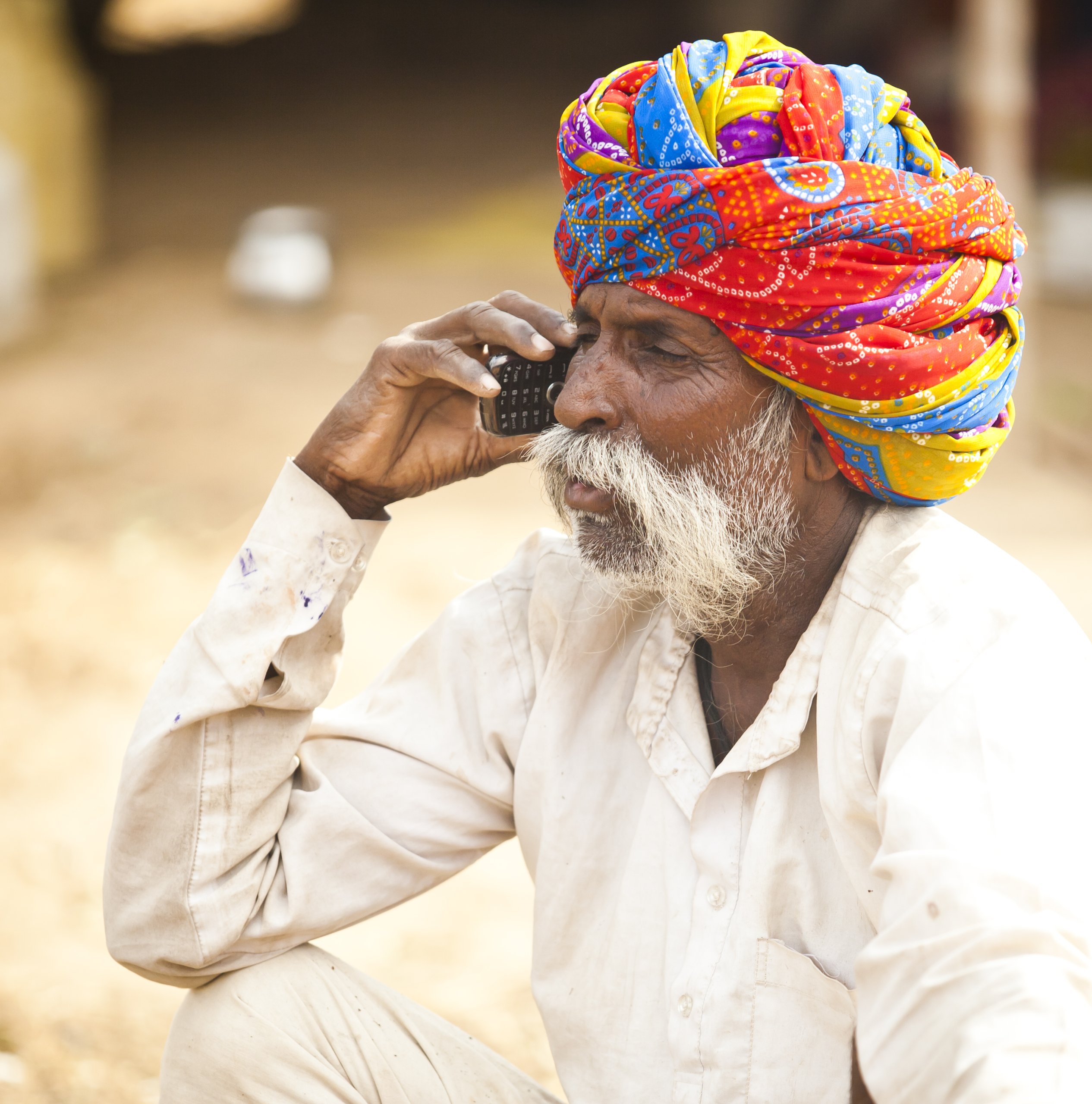 An elderly man with a white beard and mustache, wearing a colorful Rajasthani turban and a light shirt, sits outdoors holding a black cellphone to his ear—bridging the digital divide as he converses. The background is blurred and earthy.