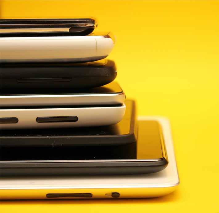 A stack of six mobile phones in varying sizes and colours, arranged horizontally on top of each other against a bright yellow background. The close-up shot highlights the sides and edges, symbolising mobile net zero efforts in technology design.