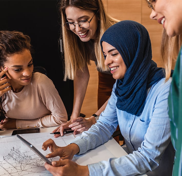 Four women collaborate around a desk, smiling and looking at a tablet. Engaged in teamwork and discussion, they review papers with diagrams—reflecting the collaborative spirit found in GSMA global, UN, and NGOs initiatives.