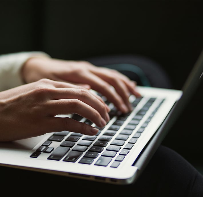 Close-up of a person's hands typing on a laptop keyboard, perhaps drafting a report on international relations. The individual wears a long-sleeved white shirt; the silver laptop’s black keys stand out against a dark, out-of-focus background.